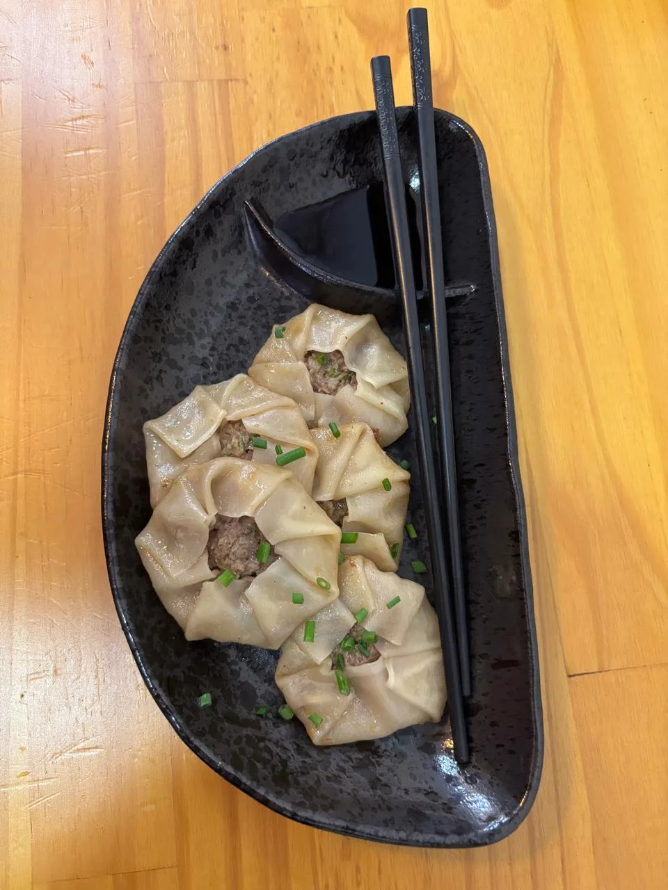 Gyozas cerradas a mano, con cebollín picado, en un plato negro junto a unos palillos